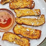 Six crispy air fryer French toast sticks dusted with powdered sugar, served on a speckled plate with warm maple syrup.