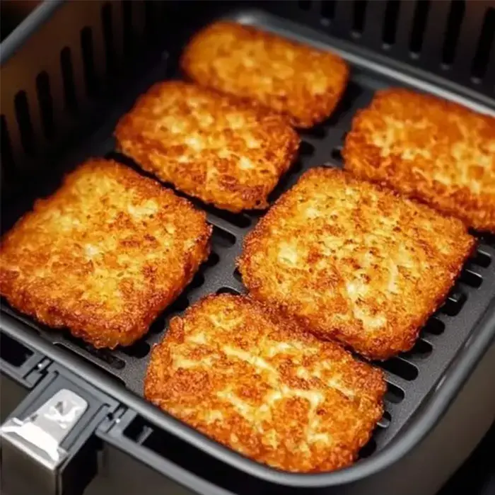 Six golden-brown, crispy frozen hash brown patties arranged in a single layer inside a black square air fryer basket.